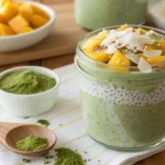 A glass jar of matcha chia pudding with coconut milk, topped with coconut flakes and mango slices.